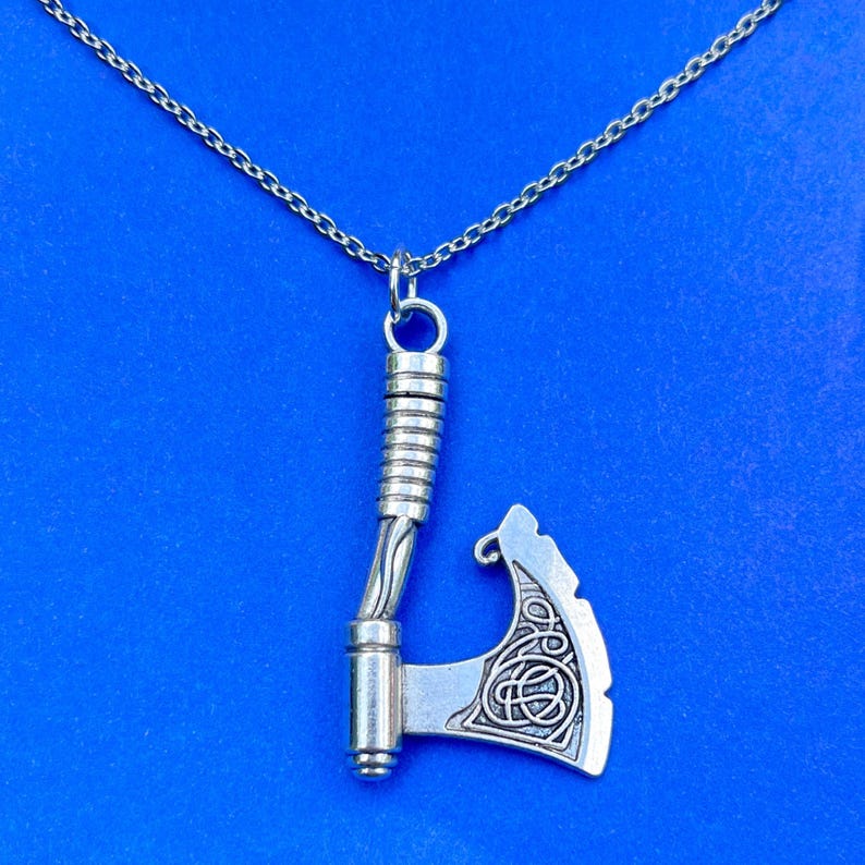 A silver Viking axe pendant necklace displayed against a vibrant blue background. The pendant features intricate Celtic knot engravings on the blade and a textured handle, giving it a detailed and historical-inspired design. The chain complements the pendant, enhancing its rugged and warrior-like aesthetic.