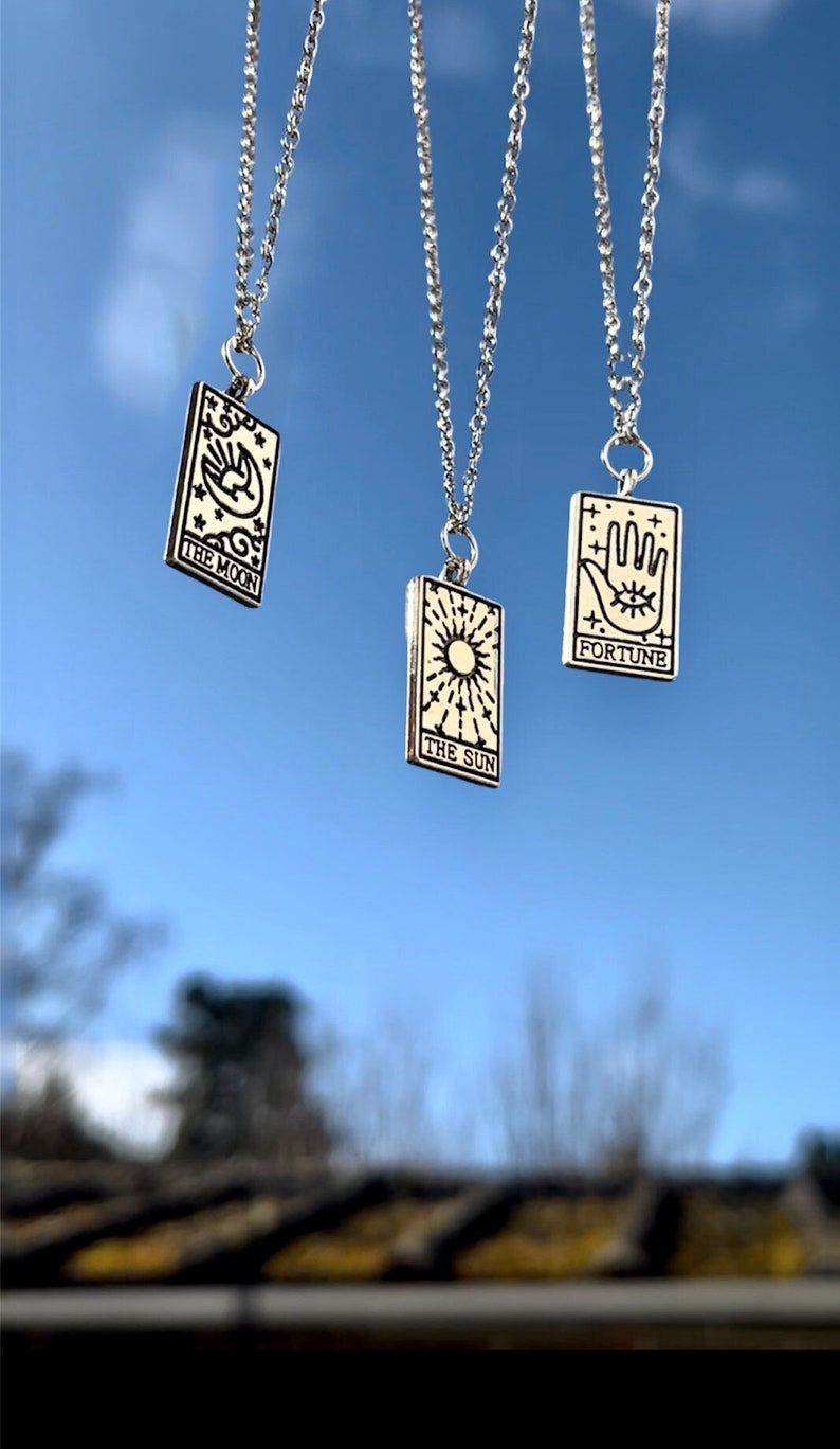 Three tarot card necklaces are pictured. From left to right they are the moon, the sun and fortune. A delicate silver chain is attached to the tarot card pendants. The pendants are rectangle shaped.