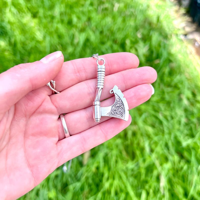 May include: Silver axe pendant necklace with intricate knotwork design. The axe is detailed with a rounded blade and a long handle.
