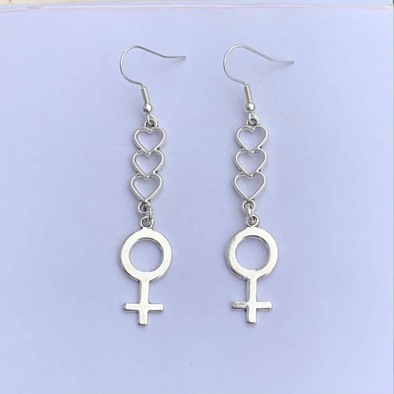 The image shows a pair of silver-colored earrings. The earrings feature a design with three connected open hearts in a vertical row at the top, followed by the female symbolat the bottom. The earrings hang from standard fishhook-style ear wires. They are a piece of silver feminist jewellery and represent women empowerment.