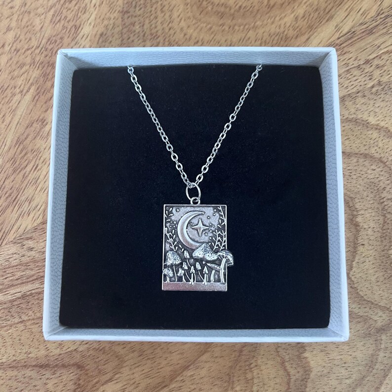 The beautiful mushroom necklace is placed against a matte black background. The detailing of the charm can be seen in more detail. The charm is silver, with some darker areas to create depth in the shapes on the charm.