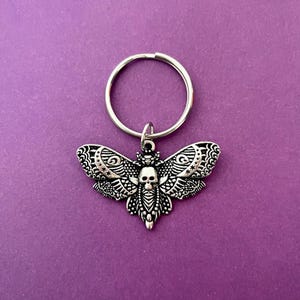 May include: A silver key chain with a detailed, ornate, silver moth charm. The moth has a skull design in the center of its body.