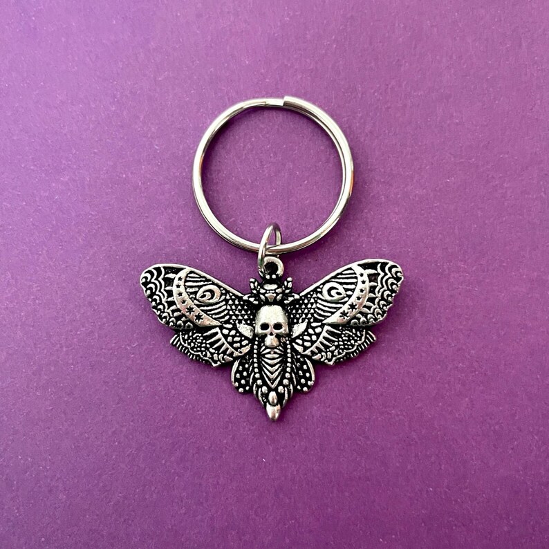 May include: A silver key chain with a detailed, ornate, silver moth charm. The moth has a skull design in the center of its body.