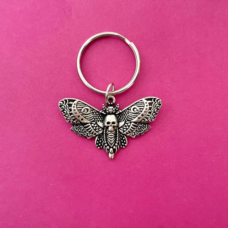 May include: A silver key chain with a detailed moth charm featuring a skull design. The moth has intricate wings and a black skull in the center.