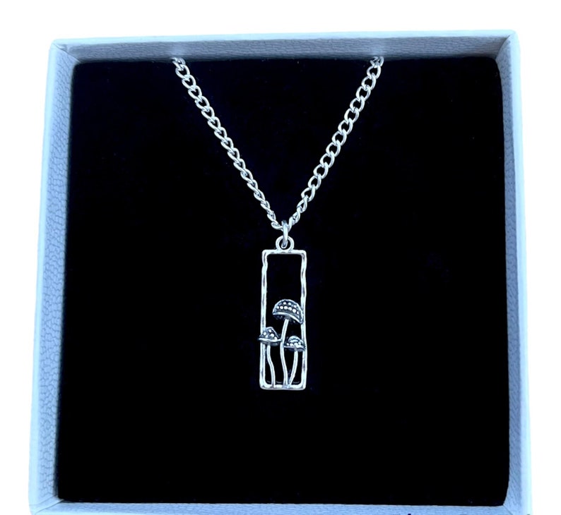 Mushroom necklace. The pendant is a rectangular outline shape. The rectangle is standing upwards. Inside this, there are three mushrooms. The largest mushroom is in the centre. The mushroom on the left is the second largest and the third smallest.