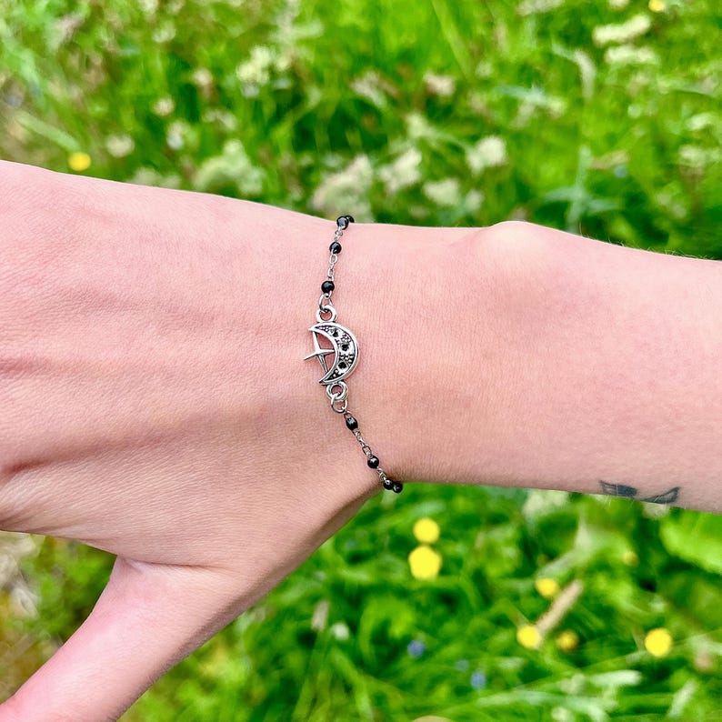 May include: A silver bracelet featuring a crescent moon charm with black accents and a star. The bracelet has a delicate chain with small black beads. The bracelet is worn on a wrist against a blurred green background.