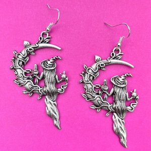 May include: A pair of silver-coloured earrings with a witch design. Each earring features a crescent moon shape with a witch figure in the centre, wearing a pointed hat and long hair. The design includes vines and leaves, with a hook for pierced ears.