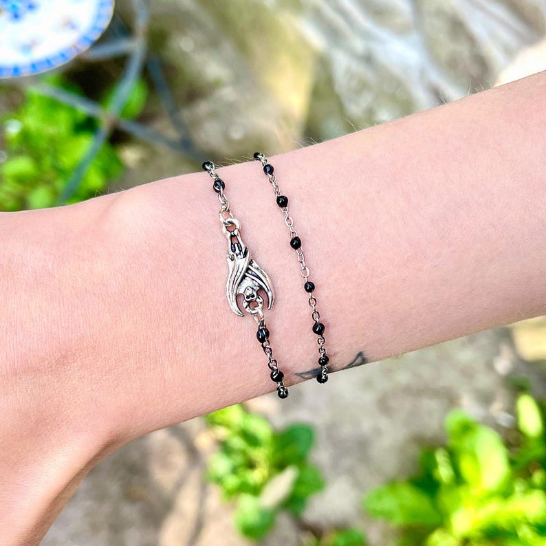 Peut inclure: Un bracelet argent&eacute; orn&eacute; d'un pendentif chauve-souris avec un motif de cr&acirc;ne. Le bracelet est compos&eacute; d'une cha&icirc;ne d&eacute;licate avec de petites perles noires. Le pendentif chauve-souris est le point central, avec des d&eacute;tails complexes.