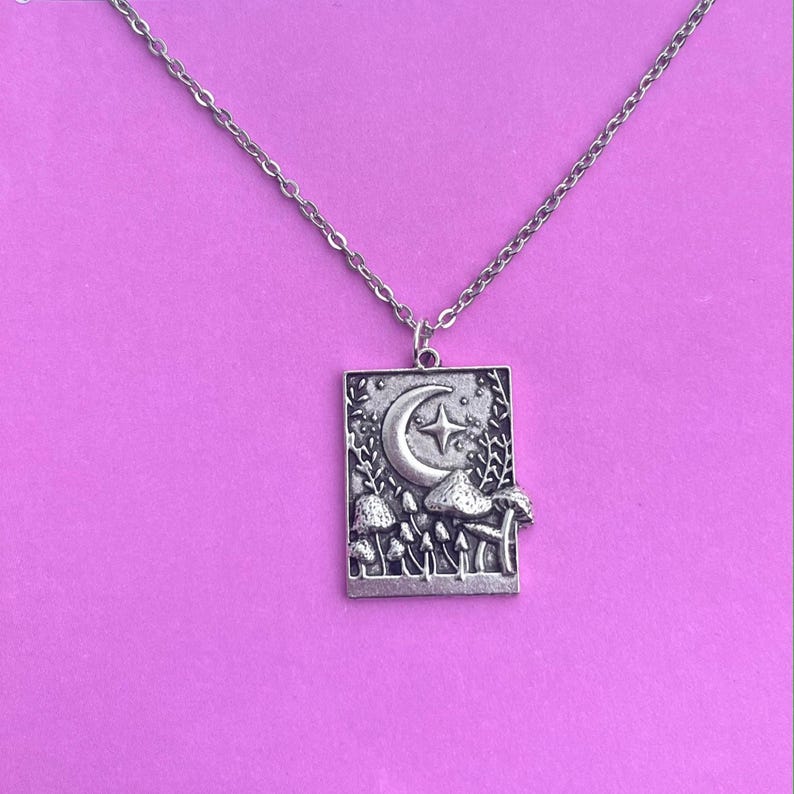 .A delicate silver chain is attached to a square-shaped charm. There is a crescent moon with a star to its right. Underneath, there are multiple mushrooms. Some mushrooms are small and some are larger. There are plants coming up the side.