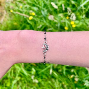May include: A silver-toned bracelet featuring a crescent moon charm with a star and black accents. The bracelet is strung with small black beads and is displayed on a wrist against a backdrop of green grass.