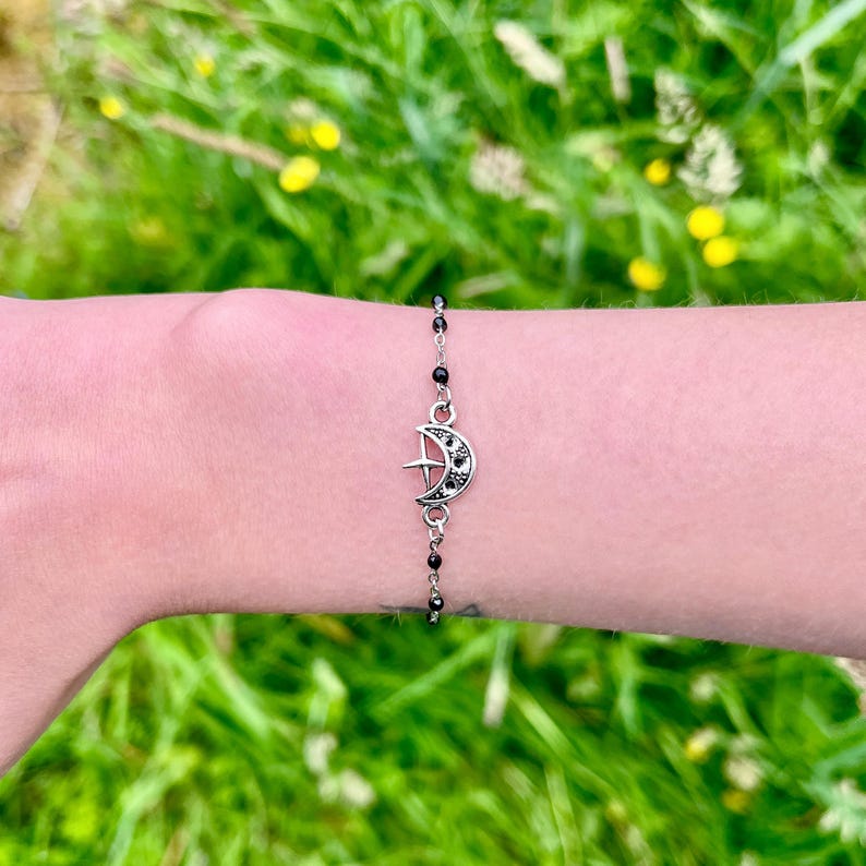 May include: A silver-toned bracelet featuring a crescent moon charm with a star and black accents. The bracelet is strung with small black beads and is displayed on a wrist against a backdrop of green grass.