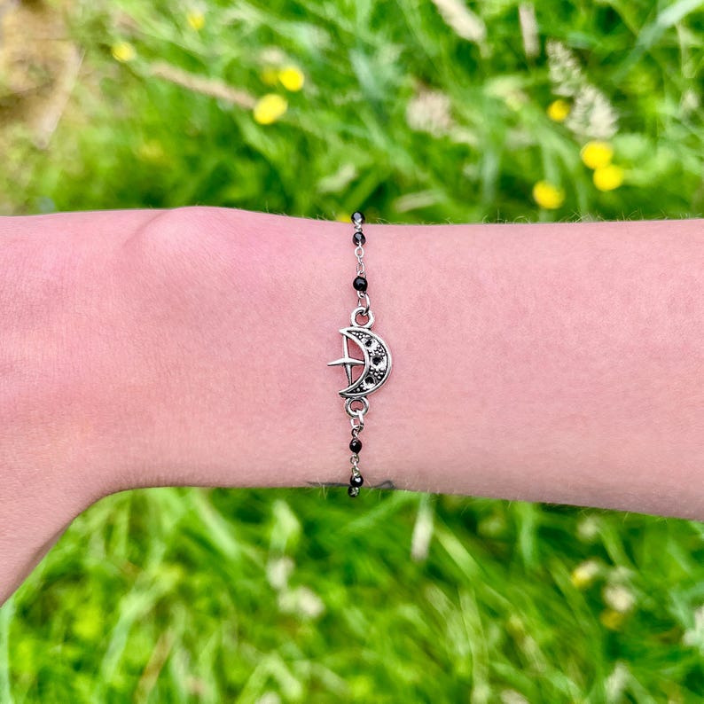 This handmade celestial bracelet features a delicate silver-tone crescent moon charm with tiny black dots and a small star detail at its center. The charm is suspended on a fine chain adorned with glossy black glass beads, adding a mystical, cosmic aesthetic. Lightweight and dainty, this moon and star bracelet offers an elegant, minimalist design perfect for layering or wearing alone. Ideal for astrology lovers, witchy fashion enthusiasts, or anyone drawn to celestial or boho jewelry styles.