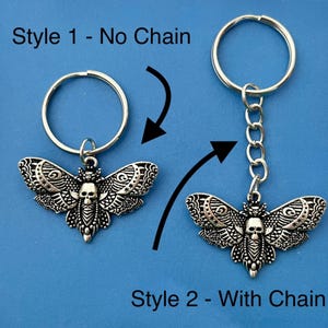 May include: Two styles of a silver key chain featuring a detailed moth with a skull design. Style 1 is a simple keychain with no chain. Style 2 has a chain attached to the keychain.