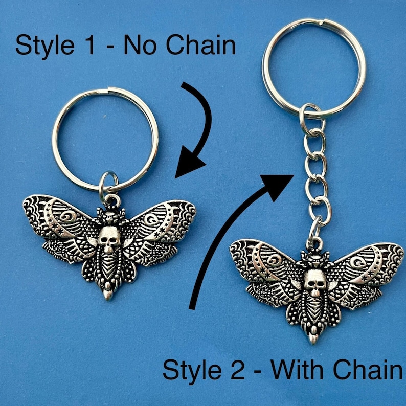 May include: Two styles of a silver key chain featuring a detailed moth with a skull design. Style 1 is a simple keychain with no chain. Style 2 has a chain attached to the keychain.