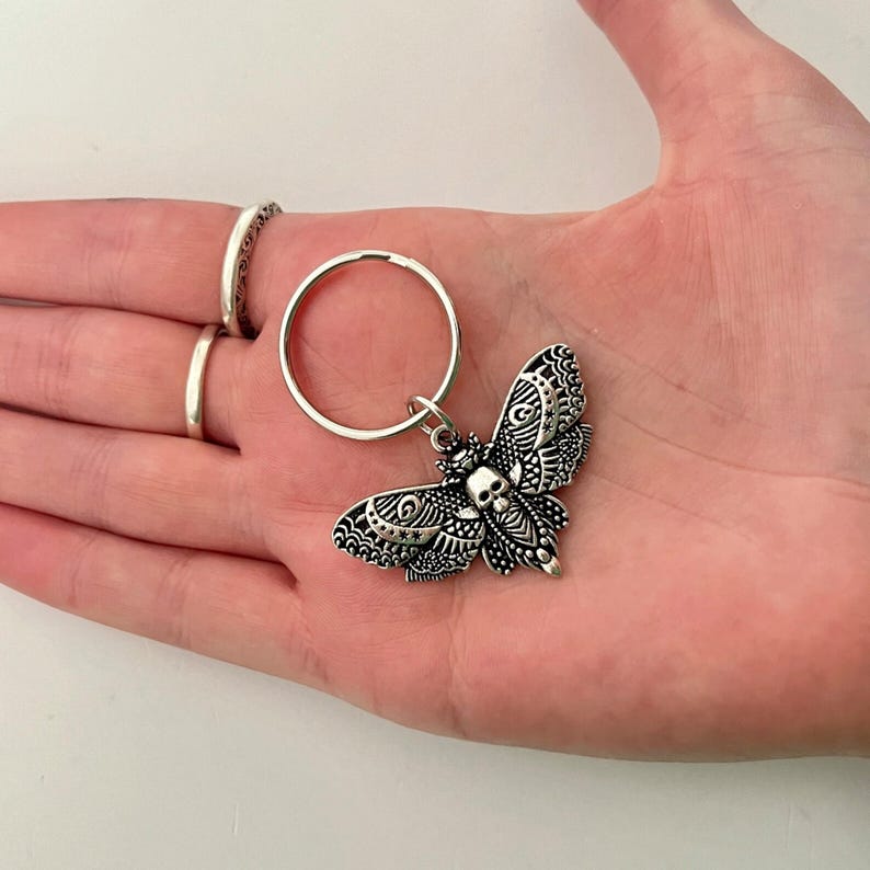 May include: Silver-toned key chain featuring a detailed, ornate moth with a skull design. The moth has intricate patterns and a black and white color scheme.