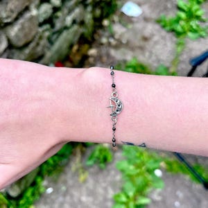 May include: Silver-toned bracelet featuring a crescent moon and star charm. The charm is embellished with small, dark stones. The bracelet has a delicate chain with small, black beads. The bracelet is worn on a wrist.
