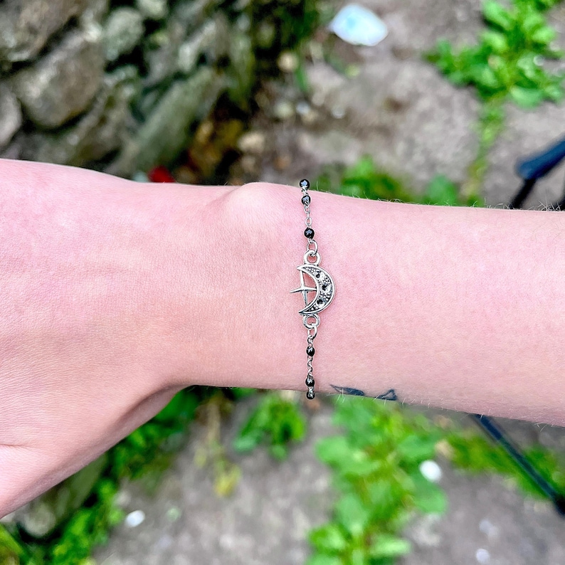May include: Silver-toned bracelet featuring a crescent moon and star charm. The charm is embellished with small, dark stones. The bracelet has a delicate chain with small, black beads. The bracelet is worn on a wrist.