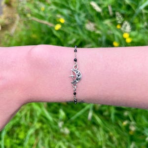 May include: Silver-toned bracelet featuring a crescent moon charm with a cross design and black accents. The bracelet is strung with small, black beads and is displayed on a person's wrist against a backdrop of green foliage.