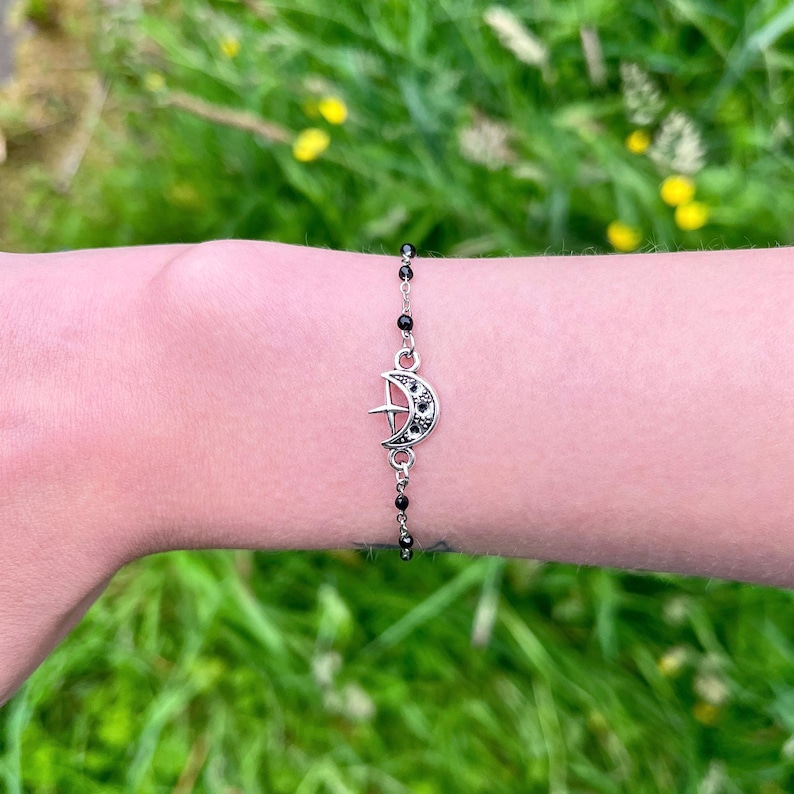 May include: Silver-toned bracelet featuring a crescent moon charm with a cross design and black accents. The bracelet is strung with small, black beads and is displayed on a person's wrist against a backdrop of green foliage.