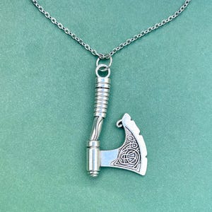 May include: A silver necklace with a detailed axe pendant. The axe has a twisted handle and a Celtic knot design on the blade.