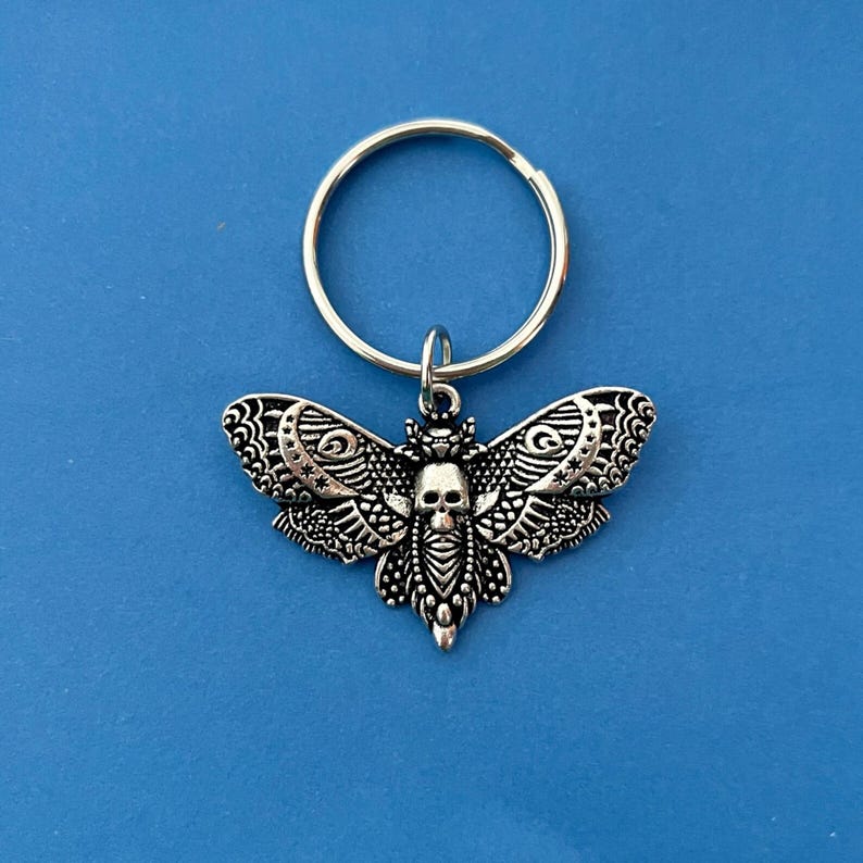 May include: A silver key chain with a detailed silver moth charm. The moth has a skull design in the center of its body.