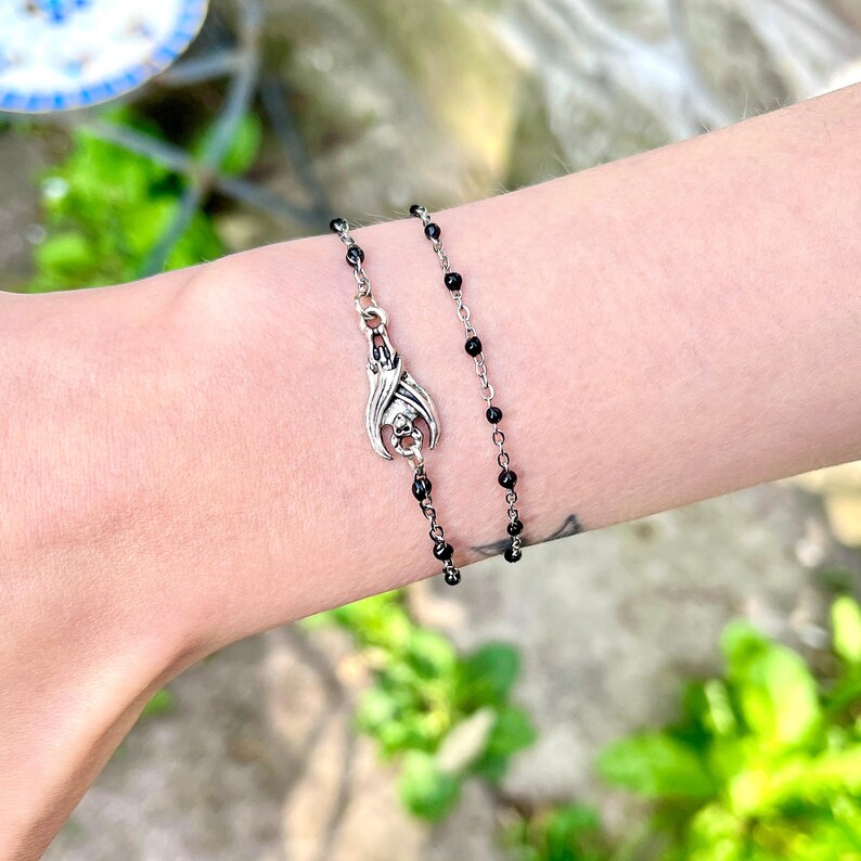 Peut inclure: Un bracelet argent&eacute; orn&eacute; d'un pendentif chauve-souris avec un motif de cr&acirc;ne et une cha&icirc;ne de perles noires. Le bracelet est port&eacute; sur un poignet, avec un tatouage visible. L'arri&egrave;re-plan est flou, avec du feuillage vert.