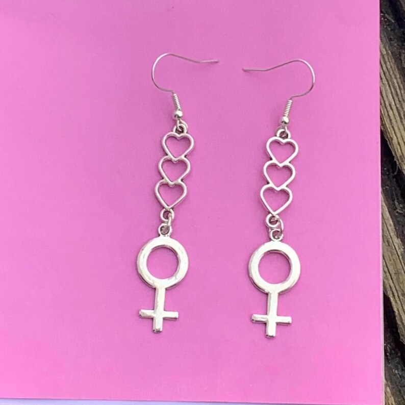 May include: Silver dangle earrings with a chain of four open heart charms and a female symbol charm at the bottom.