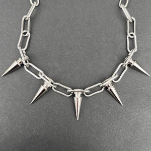 Puede incluir: Collar de cadena plateada con cinco p&uacute;as c&oacute;nicas. El collar presenta una serie de eslabones interconectados, con las p&uacute;as espaciadas uniformemente a lo largo de la parte inferior. Las p&uacute;as son met&aacute;licas y puntiagudas, a&ntilde;adiendo una est&eacute;tica audaz y vanguardista.