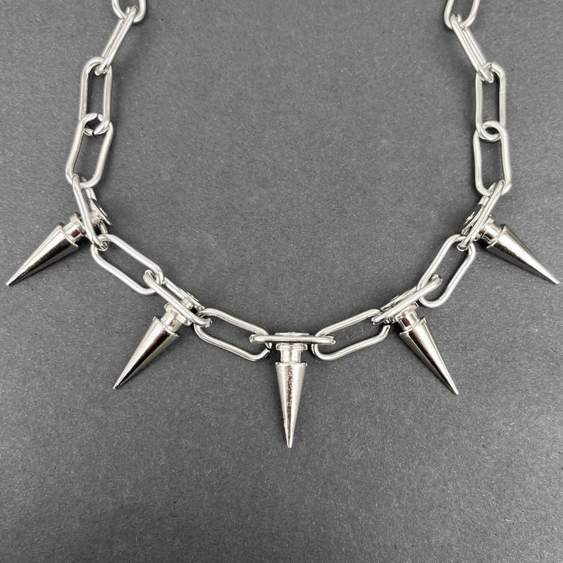 Puede incluir: Collar de cadena plateada con cinco p&uacute;as c&oacute;nicas. El collar presenta una serie de eslabones interconectados, con las p&uacute;as espaciadas uniformemente a lo largo de la parte inferior. Las p&uacute;as son met&aacute;licas y puntiagudas, a&ntilde;adiendo una est&eacute;tica audaz y vanguardista.