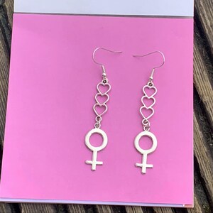 May include: Silver dangle earrings with a female symbol charm and three stacked heart charms.