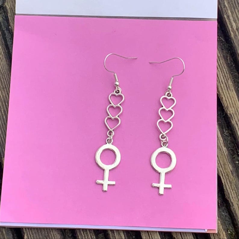 May include: Silver dangle earrings with a female symbol charm and three stacked heart charms.