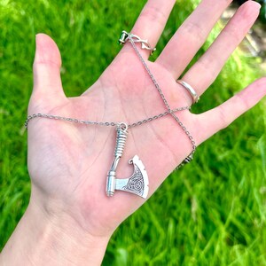 May include: A silver necklace with a detailed axe pendant. The axe has a Celtic knot design and is hanging from a silver chain.