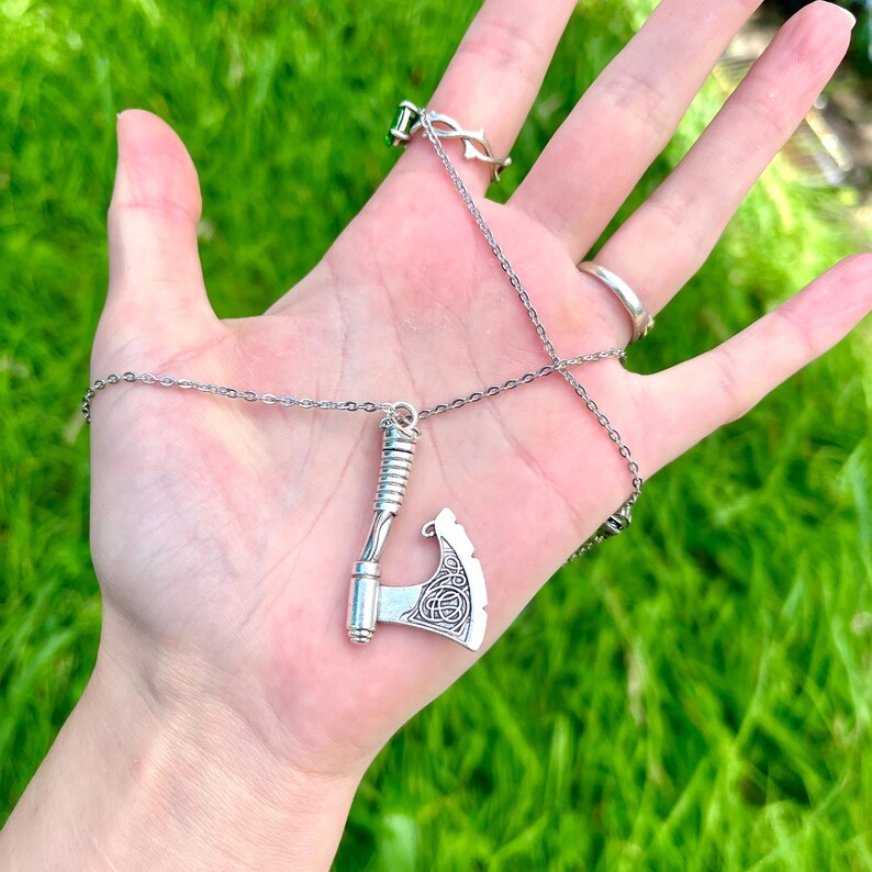 May include: A silver necklace with a detailed axe pendant. The axe has a Celtic knot design and is hanging from a silver chain.