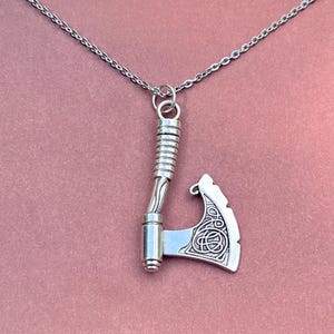 May include: A silver necklace with a detailed axe pendant. The axe has a twisted handle and a decorative design on the blade.