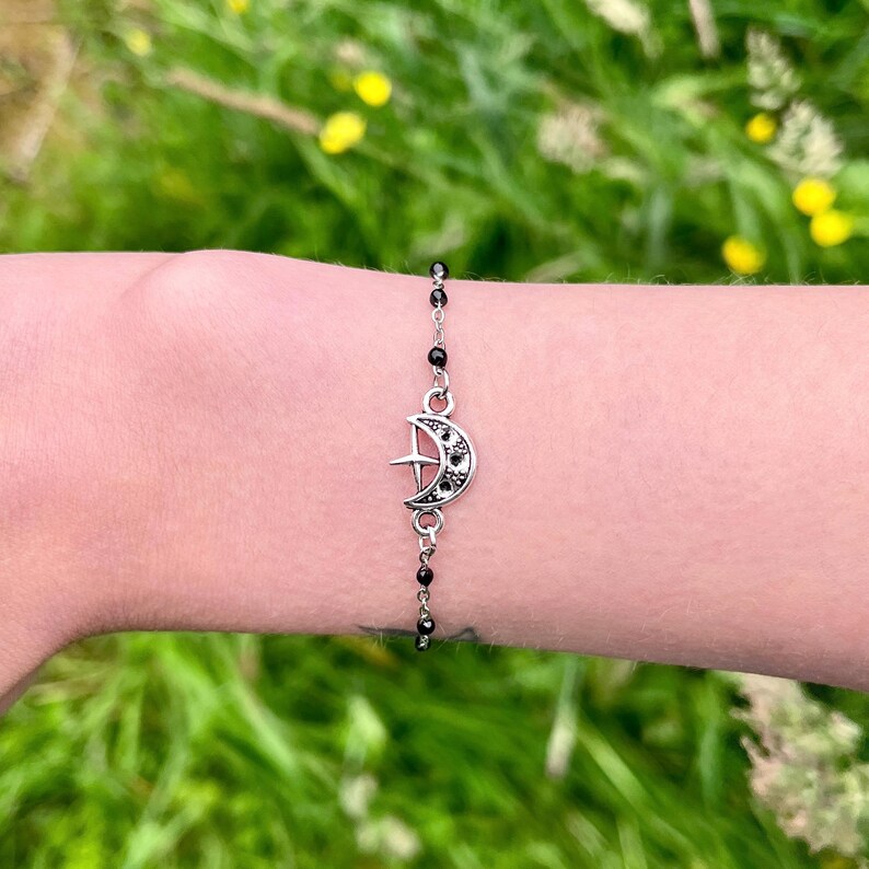 May include: Silver-toned bracelet featuring a crescent moon and star charm with black accents. The bracelet has a delicate chain with small black beads. The charm is detailed with black stones, creating a celestial-themed accessory.