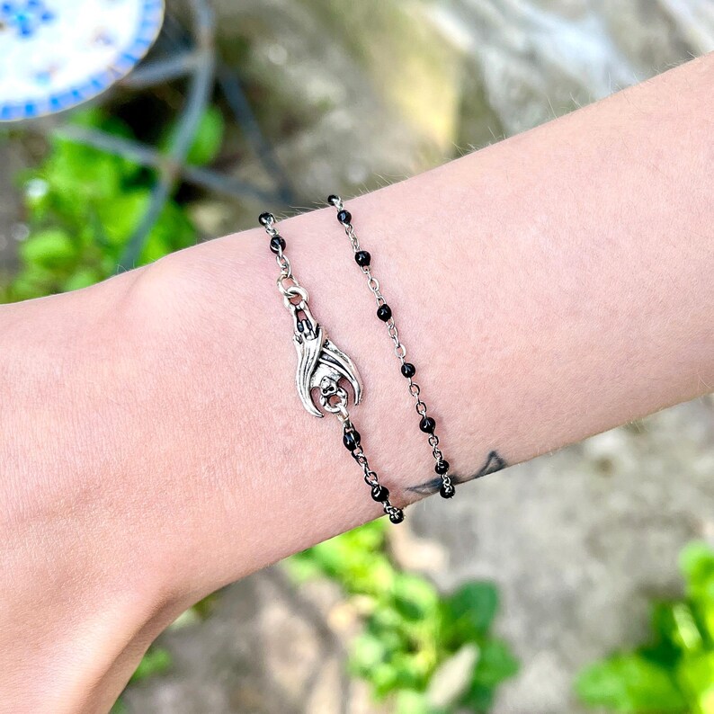 Peut inclure: Un bracelet &agrave; breloques chauve-souris argent&eacute; avec des perles noires. Le charm chauve-souris est orn&eacute; d'ailes et d'un cr&acirc;ne. Le bracelet est port&eacute; sur un poignet, mettant en valeur les bijoux gothiques.