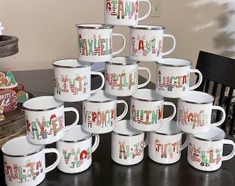 May include: A pyramid stack of white enamel mugs with silver rims, each featuring a unique name printed in colorful, festive holiday-themed lettering. The mugs are arranged on a dark surface, showcasing various personalized names in red, green, and striped patterns.