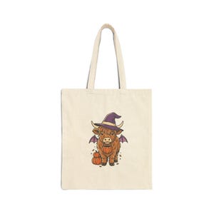 Peut inclure: Un sac fourre-tout en toile beige orné d'un motif central représentant une vache Highland brune portant un chapeau de sorcière violet et de petites ailes de chauve-souris. La vache tient un petit seau citrouille orange avec deux citrouilles au sol.