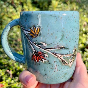 May include: A handcrafted ceramic mug in a speckled teal glaze. The mug features a floral design with yellow, red, and green accents. The handle is a matching teal color. The mug is held in a hand, with a blurred green background.
