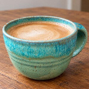 May include: A ceramic mug with a speckled finish in shades of turquoise and seafoam green, featuring a wide, rounded body and a sturdy handle. The mug is filled to the brim with a light brown coffee beverage topped with a layer of creamy foam.