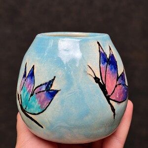May include: A small, round, light blue ceramic vessel with a glossy finish. The exterior features two hand-painted butterfly illustrations outlined in black, with one butterfly displaying teal and purple wings and the other showing pink and purple wings.