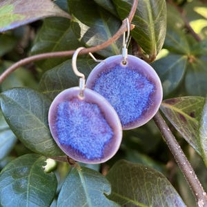 May include: A pair of handmade ceramic earrings. Each earring is a round disc with a light purple border and a vibrant blue center. The earrings hang from silver-colored hooks, set against a backdrop of green leaves and branches.