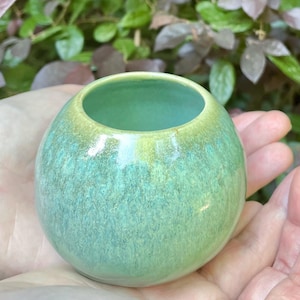 May include: A small, round ceramic vase with a wide opening. The vase has a gradient of colors, transitioning from a light green at the top to a deeper teal at the base. The surface has a textured, speckled appearance, and the vase is held in open hands.