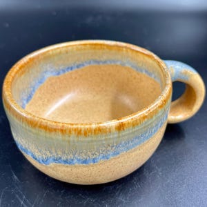 May include: A ceramic mug with a speckled tan base and a smooth, glossy finish. The rim and upper body feature a decorative band of dripped blue glaze that transitions into a warm, honey-colored brown tone around the top edge and handle.