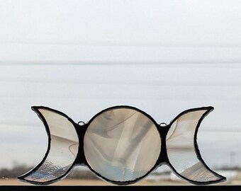 May include: Triple moon stained glass window hanging with a white background. The moon phases are represented in the stained glass.
