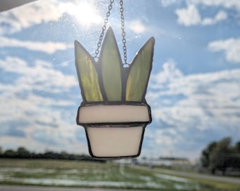 May include: A stained glass window hanging ornament featuring a potted plant with green leaves. The plant is in a white pot with a black outline. The ornament is hanging from a silver chain.