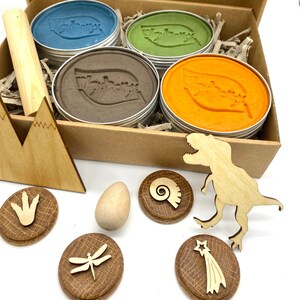 K&ouml;nnte beinhalten: Ein Set mit vier bunten Knetmassen in Metalldosen, einem Holz-Nudelholz und einem Satz Holzspielzeug zum Thema Dinosaurier, darunter ein Dinosaurier, ein Fu&szlig;abdruck, ein Fossil, eine Libelle und ein Sternschnuppe.