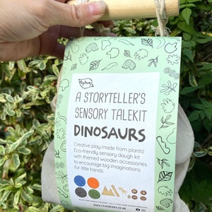 K&ouml;nnte beinhalten: Eine wei&szlig;e Tasche mit einem gr&uuml;nen Etikett, auf dem "A Storyteller's Sensory Talekit Dinosaurs" steht. Das Etikett hat einen gr&uuml;nen Rand mit einem Muster aus Bl&auml;ttern und anderen Formen. Der Text "Creative play made from plants! Eco-friendly sensory dough kit with themed wooden accessories to encourage big imaginations for little hands." steht auf dem Etikett. Die Tasche h&auml;ngt an einem Holzstab.
