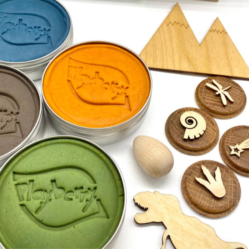 K&ouml;nnte beinhalten: Vier Dosen Playberry-Knete in Gr&uuml;n, Orange, Braun und Blau. Die Dosen sind umgeben von h&ouml;lzernen Dinosaurierspielzeug, darunter ein T-Rex, ein Berg, ein Fossil, eine Libelle, ein Stern und ein Ei.