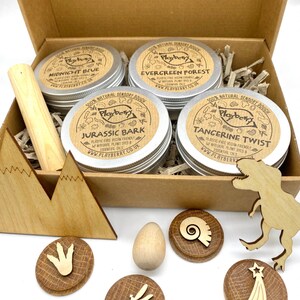 K&ouml;nnte beinhalten: Ein Set mit vier kleinen, silbernen Dosen mit nat&uuml;rlichem sensorischem Teig, beschriftet mit "Midnight Blue", "Evergreen Forest", "Jurassic Bark" und "Tangerine Twist". Die Dosen befinden sich in einer braunen Pappbox mit Holz-Dinosaurierspielzeug und einem Holzei.